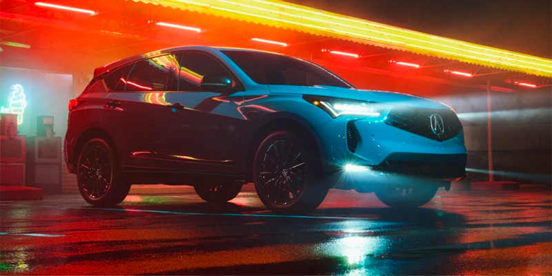A blue Acura RDX parked under neon lights at night, with a reflective wet surface adding to the dramatic lighting.