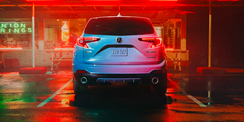 The rear view of a light blue Acura RDX parked under bright neon lights.
