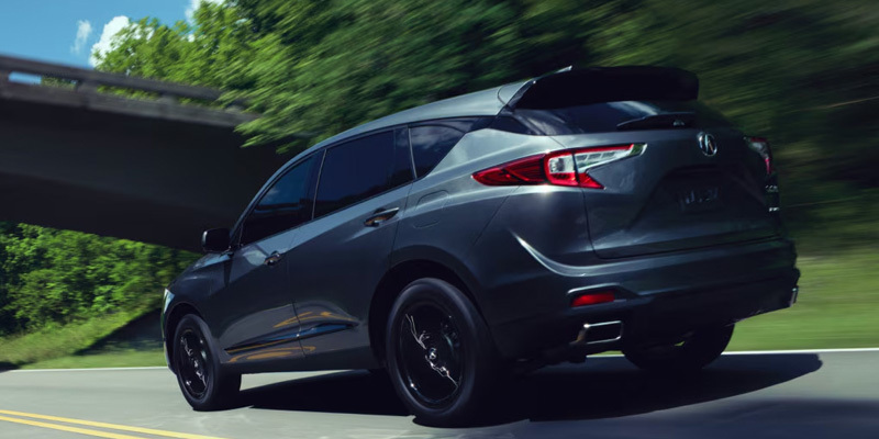 Gray Acura RDX SUV driving on a tree-lined road beneath an overpass.
