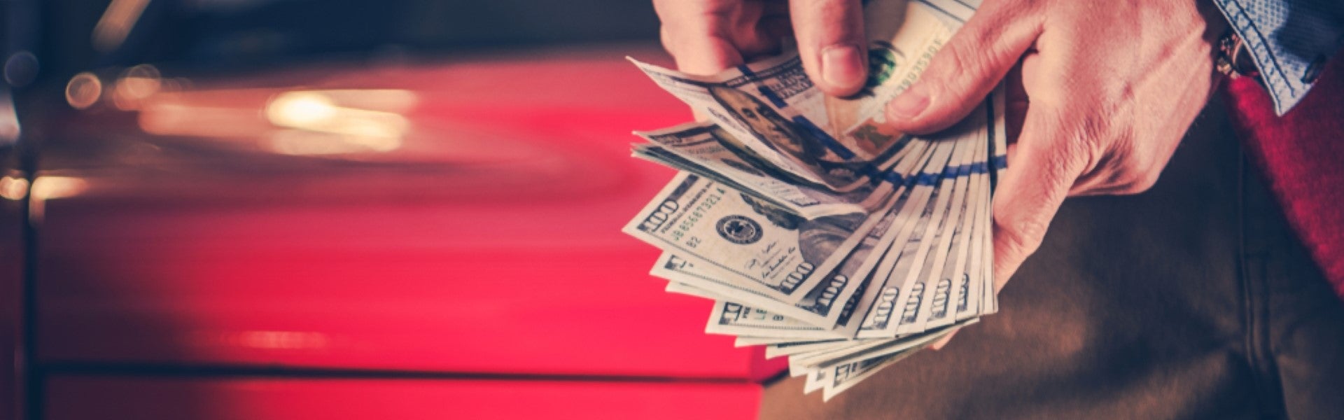 A man's hands count out several hundred dollars in front of a red car