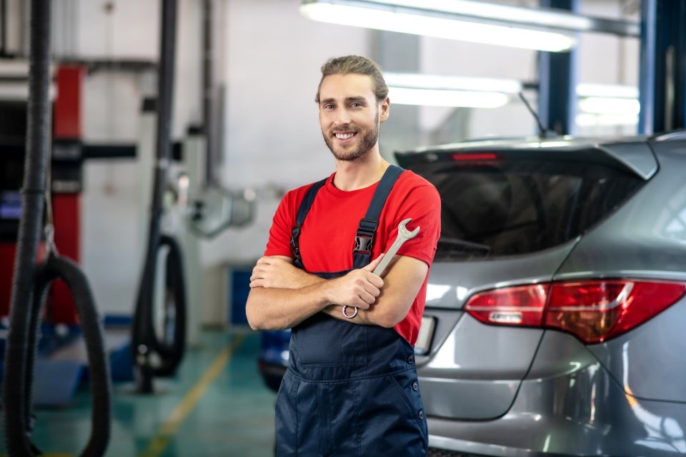 A smiling male auto mechanic holding a wrench and standing in front of a car.